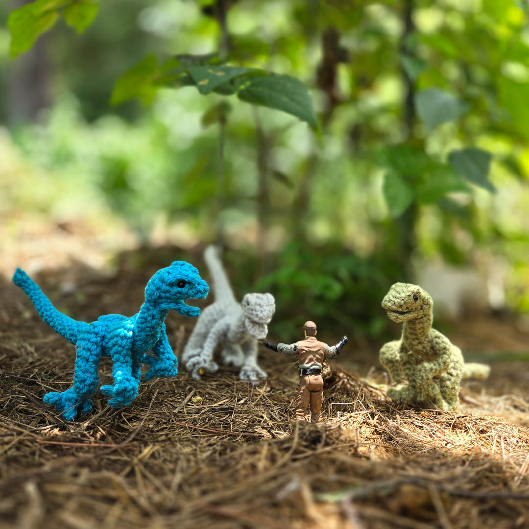 Three crochet dinosaurs clustered around a toy man with his hands raised.