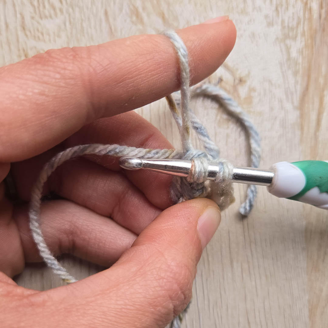 A crochet loop stitch in grey yarn wrapped around a finger, with a crochet hook poised nearby.