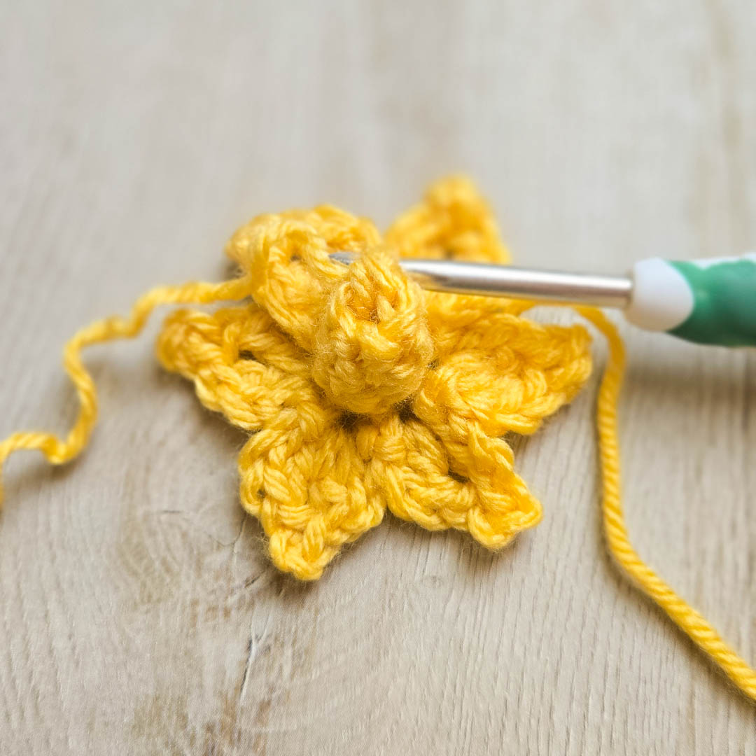 A yellow crochet flower in the works with a crochet laying on the petals.