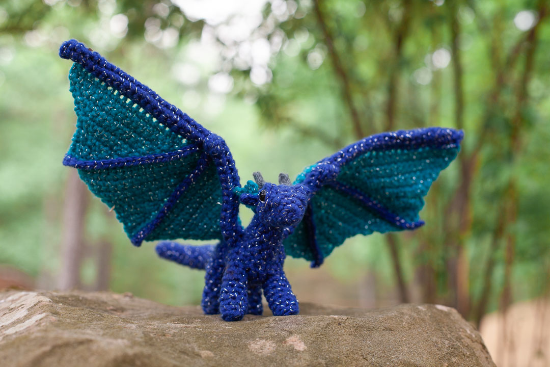 A blue crochet dragon on a rock with forest in the background.