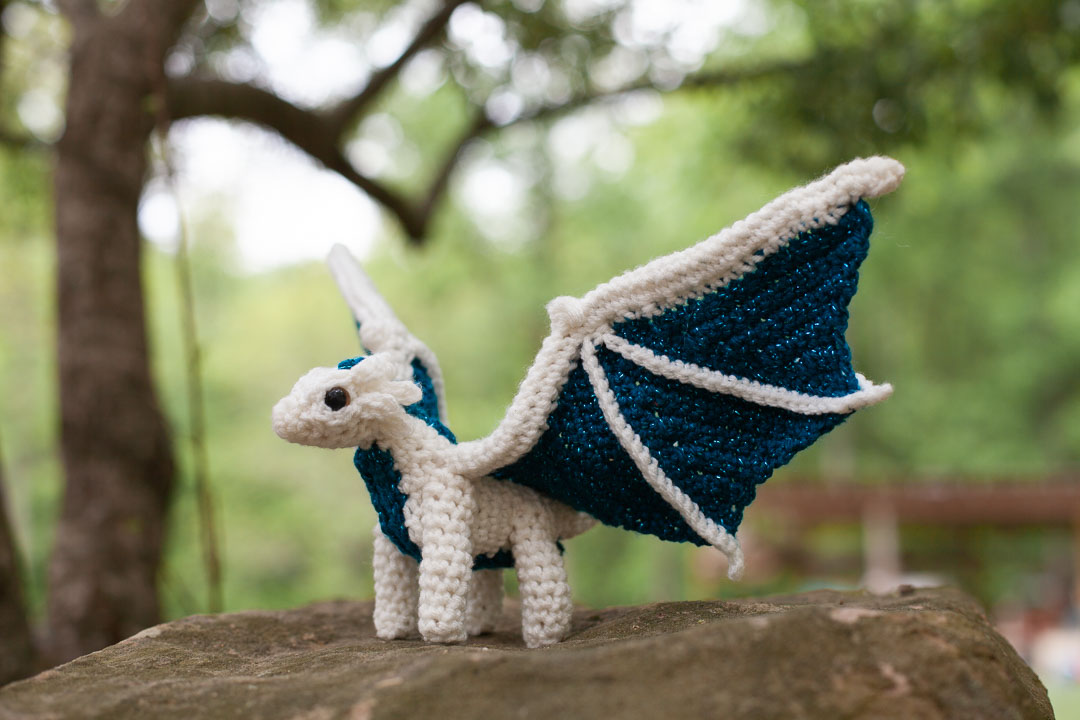 a white and blue crochet dragon perched on a rock.